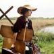 Einzelbild aus dem Film "Paula". Schauspielerin Carla Juri als Paula Modersohn-Becker ist vor einem Feld zu sehen und trägt eine Bildermappe so wie Koffer bei sich.