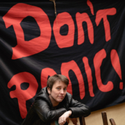 Eine junge Frau lehnt an einem Stuhl. Im Hintergund hängt eine Flagge mit Aufschrift "Don't panic!"